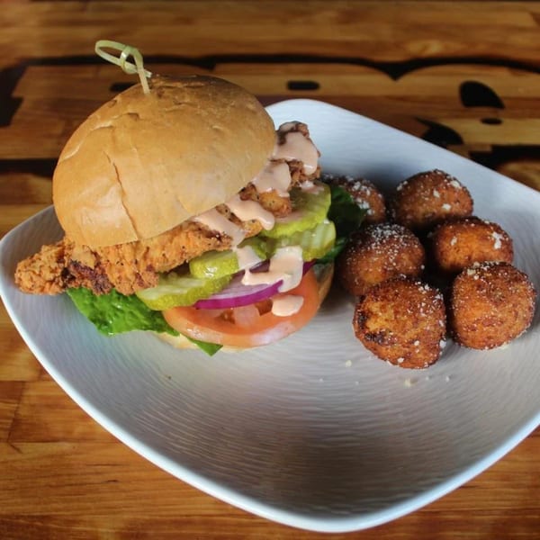 Crispy chicken sandwich with lettuce, tomato, pickles, red onion, and sauce, served with hushpuppies.