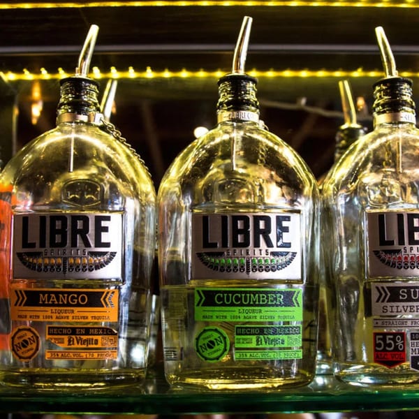 Three bottles of Libre Spirits tequila, mango, cucumber, and silver, on a lit shelf.