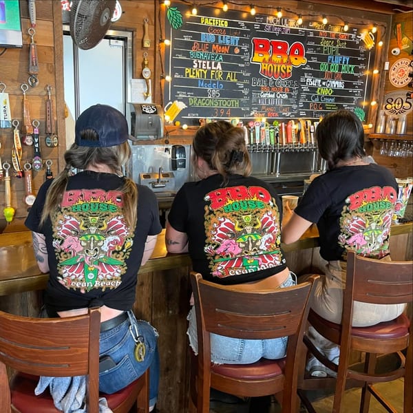 Three people wearing BBQ House shirts sit at a bar with a large chalkboard menu and beer taps.
