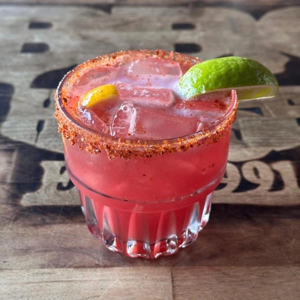 A vibrant red cocktail with ice, a chili-lime rim, and garnished with lime and lemon wedges.